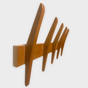 May include: A mid-century modern wooden coat rack with six angled hooks. The rack is made of light brown wood and is mounted on a rectangular base. The hooks are evenly spaced and have a unique, stylized design.
