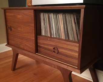 Può includere: Una console multimediale in legno scuro con un'anta scorrevole a sinistra e un vano portaoggetti aperto a destra riempito di dischi in vinile. L'unità presenta un cassetto sotto il vano, gambe affusolate e maniglie circolari in legno.