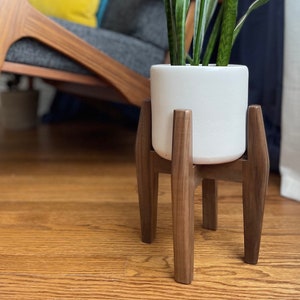 May include: A white ceramic planter with a green plant sits on a mid-century modern wooden plant stand. The stand has a dark brown finish and a tripod-like design. The background includes a wooden floor and a gray chair.