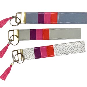 May include: Three keychains with colorful stripes and pink tassels. The keychains are made of fabric and have a silver or gold metal ring.