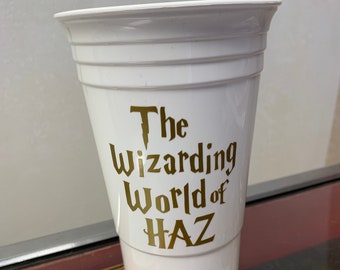 May include: White plastic cup with gold lettering that reads "The Wizarding World of HAZ".