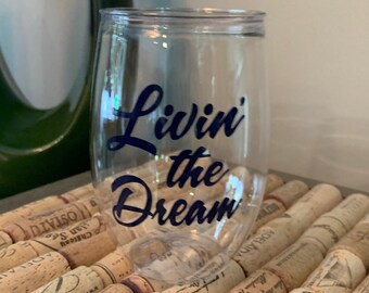 May include: A clear plastic wine glass with a blue vinyl decal that reads "Livin' the Dream". The glass is sitting on a surface covered in cork wine bottle stoppers.