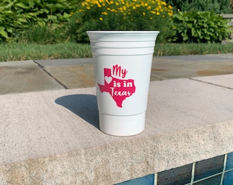 May include: White plastic cup with a pink heart and the text "My heart is in Texas" printed on it. The cup is sitting on a white surface near a pool.