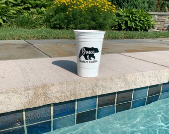 May include: White plastic cup with a black bear silhouette and the text "Bruce Family Cabin" printed on it. The cup is sitting on the edge of a swimming pool.
