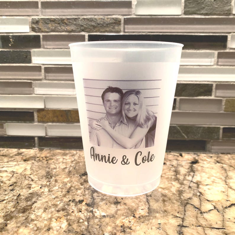 May include: A white plastic cup with a black and white photo of a couple in a mugshot style frame. The text "Annie & Cole" is printed below the photo.