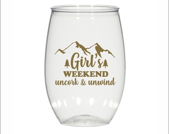 May include: A clear plastic wine glass with a gold mountain and tree design and the text "Girl's Weekend uncork & unwind".