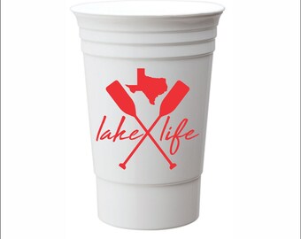 May include: White plastic cup with a red graphic of crossed paddles and the state of Texas with the text "lake life" printed on it.