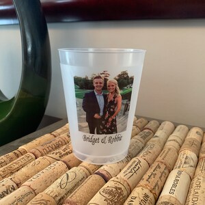 May include: A clear plastic cup with a photo of a couple printed on it. The photo shows a man and a woman standing in front of a green background. The text "Bridget & Robbie" is printed below the photo.