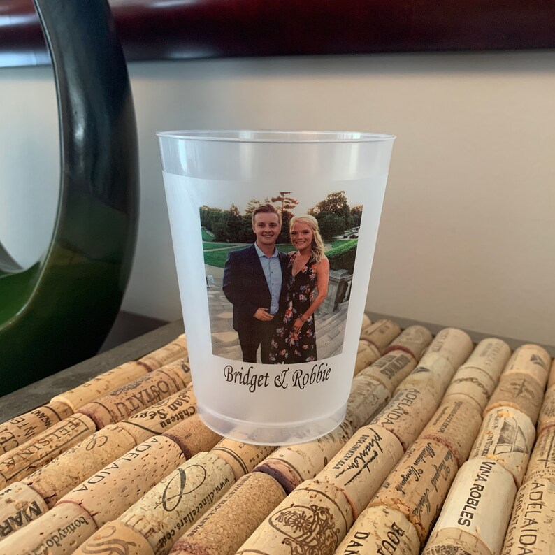 May include: A clear plastic cup with a photo of a couple printed on it. The photo shows a man and a woman standing in front of a green background. The text "Bridget & Robbie" is printed below the photo.