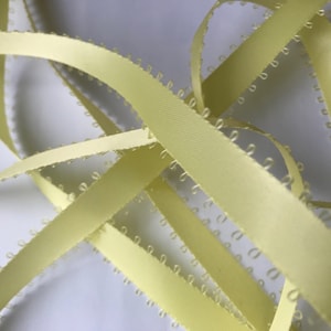May include: A close-up of yellow satin ribbon with a scalloped edge. The ribbon is folded and twisted, creating a decorative pattern.