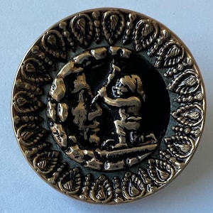 May include: A round, antique-style button with a detailed relief design. The central image depicts a figure carving a rock, surrounded by a dark background. The button has a decorative border with a leaf pattern and a metallic gold finish.