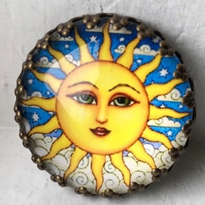 May include: A round, decorative pin featuring a smiling sun with yellow rays, green eyes, and a red mouth. The background is blue with white clouds and stars. The pin has a bronze-colored, ornate metal border.