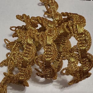 May include: Close-up of a collection of gold-colored, woven decorative trim. The trim is intricately designed with a textured, braided appearance. The metallic material reflects light, creating a shimmering effect. The trim is likely used for embellishing clothing or crafts.