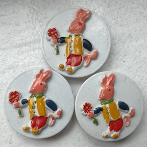 May include: Three white, circular buttons with a raised design of a pink rabbit holding a red flower. The rabbit wears a yellow vest, red pants, and a green scarf. The buttons are likely for sewing or crafts.
