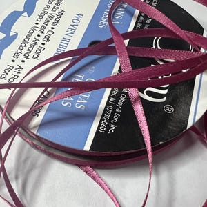 May include: A spool of woven ribbon in a deep pink hue, with strands cascading outwards. The label reads "WOVEN RIBBON" and includes text in multiple languages. Ideal for apparel, craft, and floral projects.
