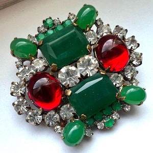 May include: A brooch featuring two large rectangular green stones, two large oval red stones, and several smaller green cabochons and clear faceted rhinestones set in a gold-tone metal frame. The design is symmetrical with a cluster of gems.