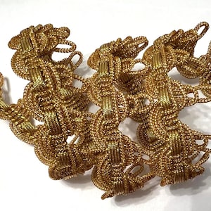 May include: Close-up of a gold-colored decorative trim. The trim is made of woven metallic threads, creating a textured, chain-like pattern. The trim is likely used for embellishing clothing or home decor items.