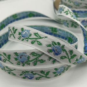 May include: White ribbon with a floral design featuring blue flowers, green leaves, and silver accents. The ribbon's edge has a mosaic pattern in shades of blue and green. This decorative ribbon is suitable for crafts and embellishments.