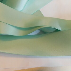 May include: A close-up of a light green satin ribbon. The ribbon is folded and arranged on a white surface.