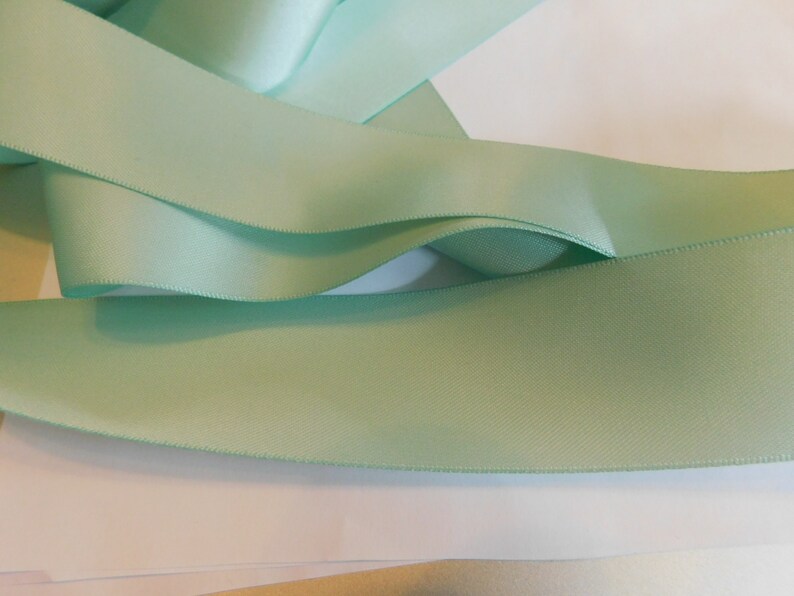May include: A close-up of a light green satin ribbon. The ribbon is folded and arranged on a white surface.