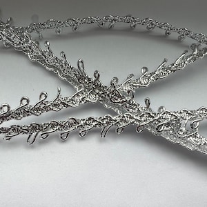 May include: Silver metallic trim with a delicate, intricate design. The trim has a woven, braided look with small loops and curves.