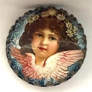 May include: A round, vintage-style image of a cherub with brown hair, a floral crown, and pink wings. The cherub's face is centered, surrounded by a blue and white floral border. The image has a slightly aged appearance.