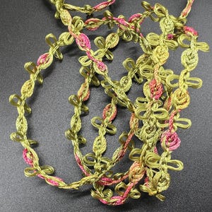 May include: Close-up of decorative trim in a looping design. The trim features a combination of olive green and pink threads, creating a textured and colorful accent. The trim is likely used for embellishing clothing or craft projects.