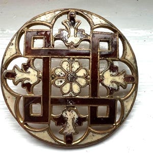 May include: A round, vintage brooch with a gold-tone metal frame. The brooch features a geometric design in dark brown and off-white enamel, with a central flower and decorative leaf accents. The design is intricate and detailed.
