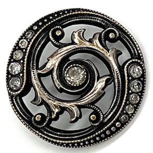 May include: A round, antique-style button with a black outer ring and a silver-colored, swirling floral design in the center. The outer ring is studded with clear, sparkling rhinestones. The button has a vintage aesthetic.