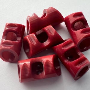 May include: A collection of six cylindrical, red beads with carved geometric designs. Each bead has a central hole and a series of curved indentations. The beads are arranged on a white surface.