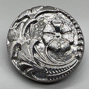 May include: A silver metal button with a floral design. The button has a raised flower in the center and intricate scrollwork around the edges.