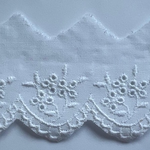 May include: White eyelet lace trim with a scalloped edge and floral embroidery. The top edge has a zig-zag design. This delicate trim is suitable for various sewing and craft projects.