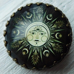 May include: Round, dark brown decorative object featuring a sun face design with radiating lines, crescent moons, and stars. The sun face is cream-colored with a smiling expression. The edge is trimmed with small, bronze-colored floral accents.