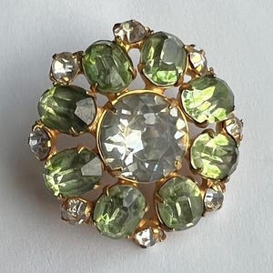 May include: A circular brooch featuring a large, central clear faceted stone surrounded by a ring of oval-shaped light green stones and smaller, clear round stones set in a gold-toned metal frame with prong settings throughout the design.