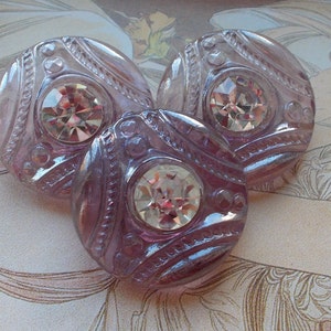 May include: Three round, lilac-coloured buttons with a central, clear, faceted jewel. The buttons have an embossed design with a beaded border. They are arranged in a triangular pattern. These buttons are likely for sewing or crafts.