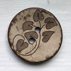May include: A round, light brown button with a dark brown rim. The button features a carved design of a plant with heart-shaped leaves and two holes for sewing. The button is made of a natural material.