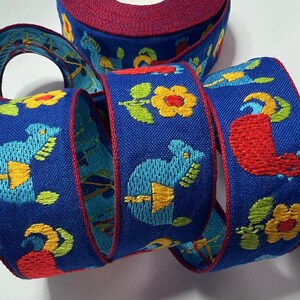 May include: A roll of blue ribbon with embroidered designs. The ribbon features repeating patterns of stylized animals, flowers, and other shapes in yellow, red, and green. The edges are trimmed with a red border.
