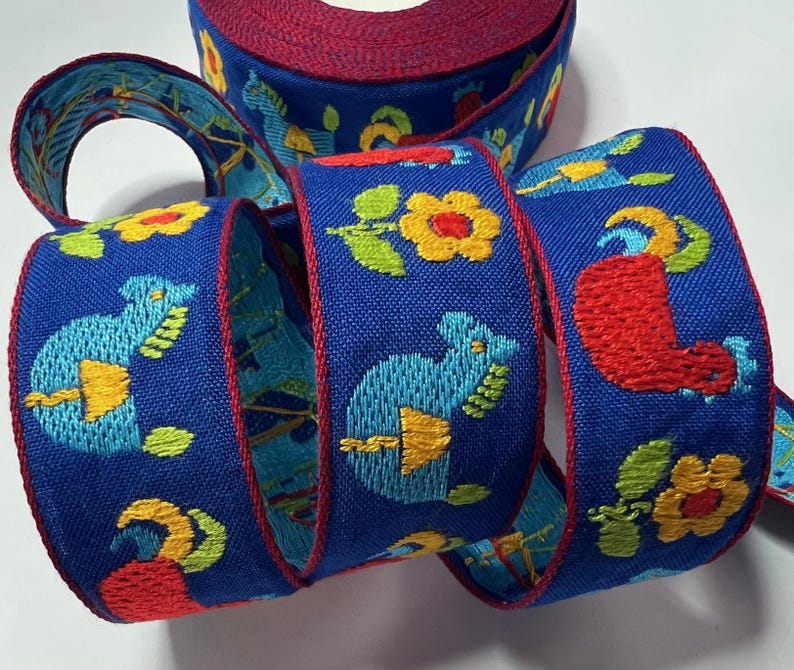May include: A roll of blue ribbon with embroidered designs. The ribbon features repeating patterns of stylized animals, flowers, and other shapes in yellow, red, and green. The edges are trimmed with a red border.