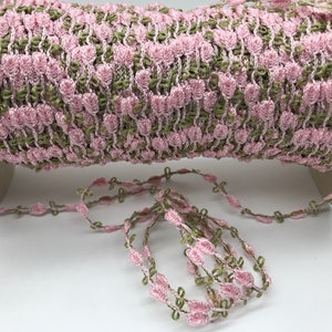 May include: A spool of pink and green floral trim. The trim features a repeating pattern of pink flowers and green leaves. A length of the trim is unspooled and lies in front of the spool.