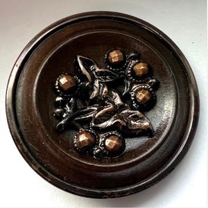 May include: A circular dark brown button featuring a central metallic relief design of leaves and stems accented with five small, faceted bronze-colored studs arranged around the perimeter of the central botanical motif.