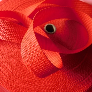 May include: Close-up of a roll of bright orange webbing. The webbing is woven with a textured pattern and is rolled up tightly.