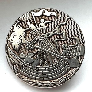 May include: A round, silver-toned button with a detailed relief design. The design features a ship with figures, a goat head, and other symbolic elements. The button has a textured background and a raised border.