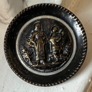 May include: A round, black button with a raised, silver-colored design. The design features two figures in relief, one holding a staff and the other holding a book. The button has a beaded edge.