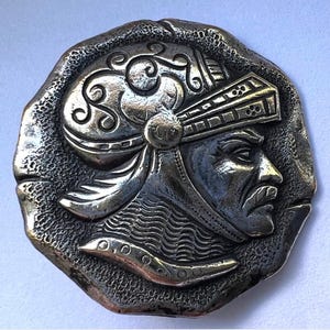 May include: A circular silver-toned metal medallion featuring a relief profile of a person wearing an ornate helmet with scrollwork and a chainmail neck guard. The background of the medallion has a textured, hammered finish with irregular edges.