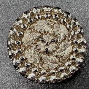 May include: A round metallic button featuring a central embossed design of swirling leaves and petals. The outer edge is decorated with a border of faceted, reflective studs that create a shimmering, geometric pattern around the center.