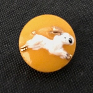 May include: A yellow button with a white rabbit jumping on it. The rabbit has black eyes and a gold outline.