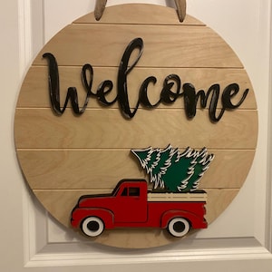 Peut inclure: Panneau de bienvenue rond en bois avec un camion rouge transportant un sapin de Noël vert. Le mot "Welcome" est en écriture noire. Le panneau est en bois clair et possède une corde de jute pour l'accrocher.