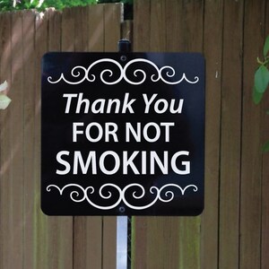 May include: A black rectangular sign with white text that reads Thank You FOR NOT SMOKING. The sign features white decorative scrollwork at the top and bottom edges and is mounted on a thin, vertical metal stake for placement in the ground.