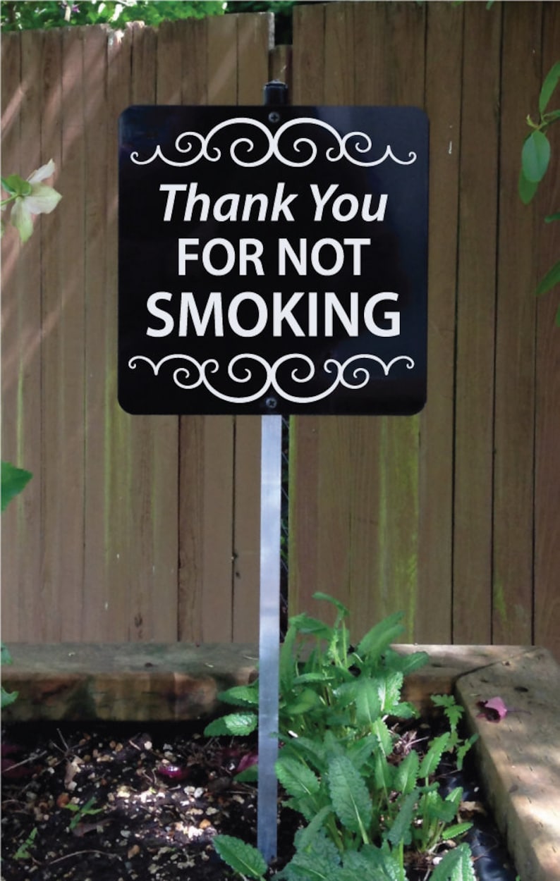 May include: A black rectangular sign with white text that reads Thank You FOR NOT SMOKING. The sign features white decorative scrollwork at the top and bottom edges and is mounted on a thin, vertical metal stake for placement in the ground.