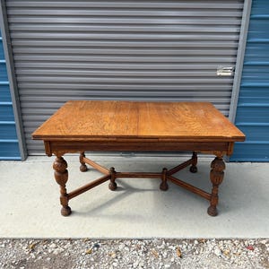 May include: A rectangular wooden dining table with a natural brown finish and visible wood grain. The table features four carved legs with decorative bulbous detailing and a central X-shaped stretcher base connecting the legs for support.